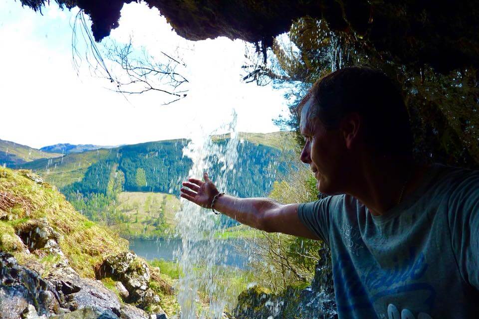 Man in cave behind waterfall. He is reaching out to touch the falling water.