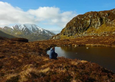 small Scottish lochan with a man sitting on the shore. A snow-capped mountain is in the background