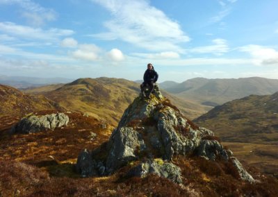 man sitting on a large rocky peak with Scottish Highland hills in the background