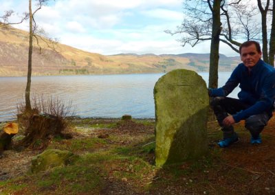 man kneeling beside a faded gravestone by a Scottish Highland loch