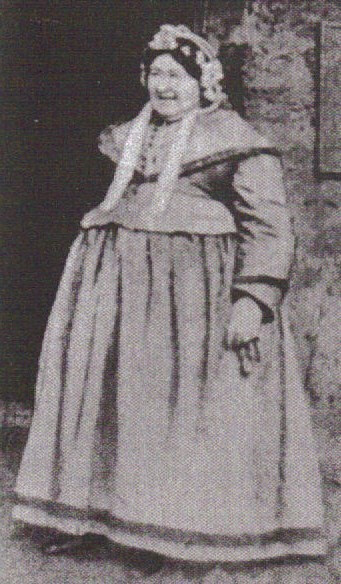 very large woman in a traditional Highland Scottish dress standing outside of a stone house