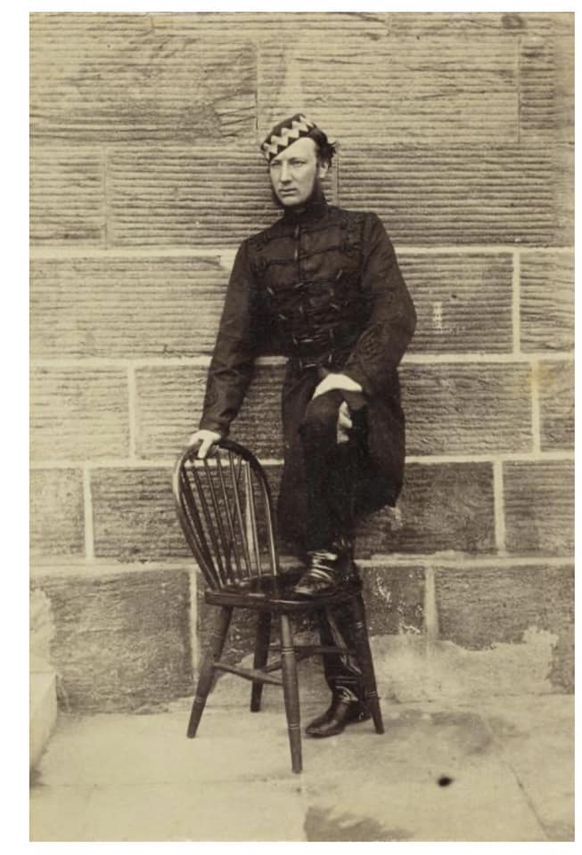 man in 19th century British military uniform leaning against a brick wall with one foot resting on a wooden chair