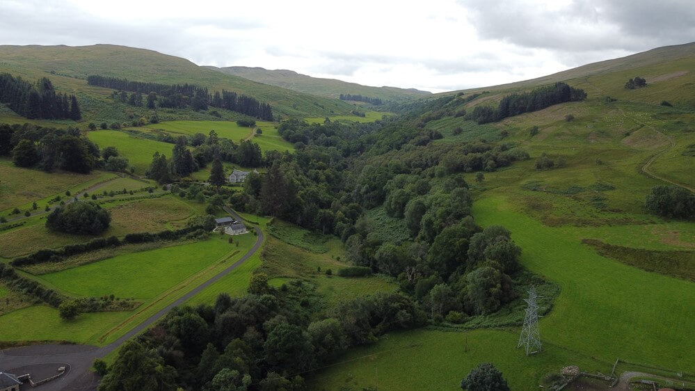 aerial view of a highland Scottish valley with lush green grass and a tree line creek winding up into the hills, beneath a mostly cloudy sky