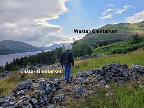 view of a Highland Scottish loch from up high on a hillside. A white-haired man in a motorcycle jacket stands on a pile of rubble looking at the view with his back to the camera