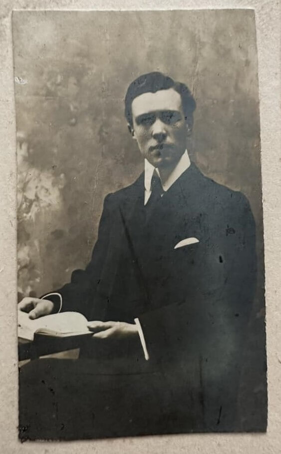 b&w photo, early 1900s, of a well-dressed man in his 30s, sitting posed at a desk, holding an open book as a prop