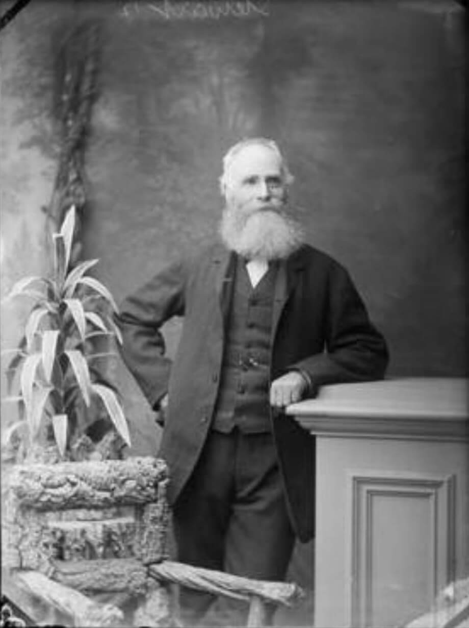 late 19th century b&w photo of an elderly man with a long beard posing in a studio wearing a suit and leaning against a desk