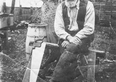 b&w photo of a man in his 60s with a grey beard, sitting on a saw horse in his yard, with his knees crossed, a saw propped by his side, and a bale of chicken wire behind him. He is smiling and the lines around his eyes are distinct.