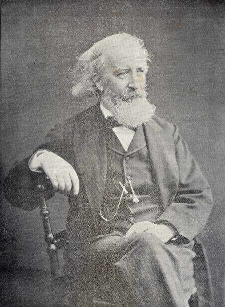 elderly man, late 1800s, in a suit, with long hair and a beard, sitting for a posed studio portrait. His right arm is draped over the back of the chair. His left hand is resting in his lap.