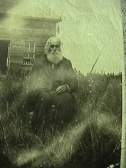 damaged photo ca. 1910 of an elderly farmer in his 70s, wearing a dark suit, sitting on a wooden chair in the long grass and weeds in front of his farm house.