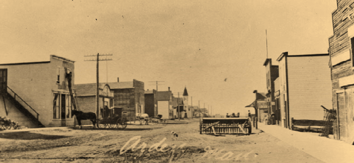 b&w image of an old prairie town, ca. 1910, showing a dirt street flanked by a handful of wooden stores and businesses. A lone horse and buggy sits tethered under a hydro pole.