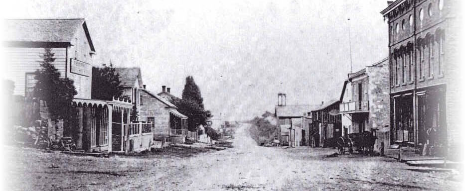 b&w image of an old pioneer town, ca. 1900, showing a dirt street flanked by a handful of wooden stores and businesses, with one or two trees on the street.