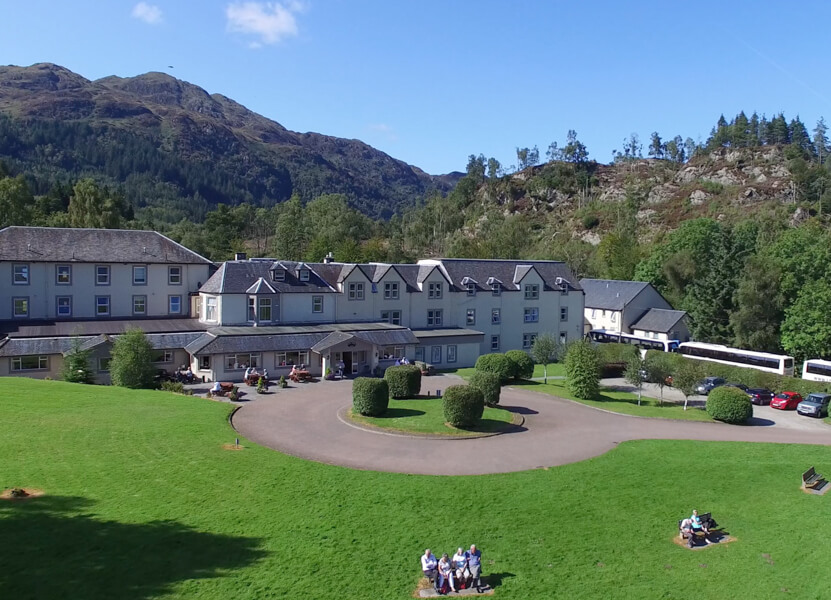Modern luxury hotel in a 19th century converted estate house in the Highlands of Scotland. The hotel is white with a rocky craig viewable behind to the right and a large mountain to the left. The sky is blue with small clouds. The foreground is a manicured lawn with garden tables and a circular driveway.