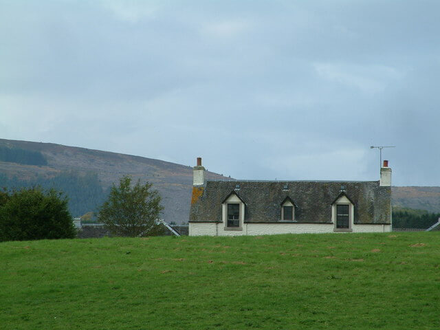 a 19th century farmhouse partially obscured from view by a low hill, under a cloudy sky, with mountain slope in the distance partially in view to the left behind the house