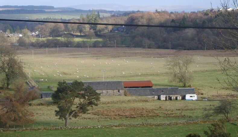 Long-distance picture of a renovated 19th century farm town There is a flat field behind the house dotted with sheep. Beyond the field are trees on gentle slopes of land.