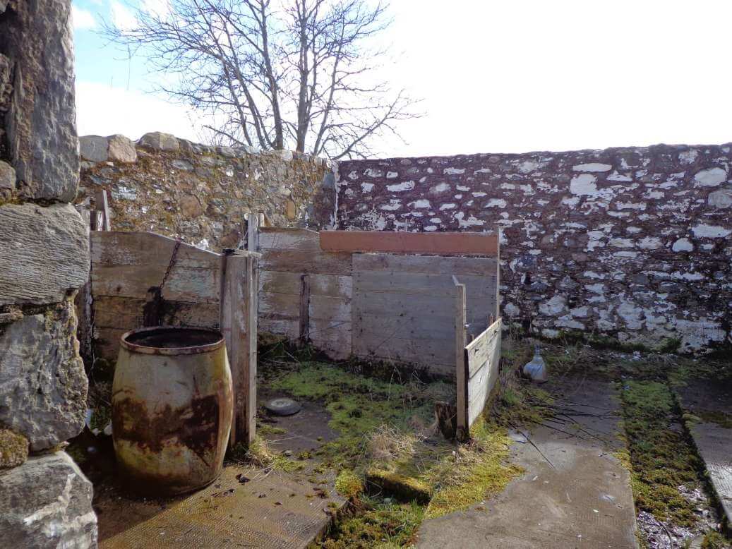 remains of a wooden animal pen within the stone walls of the ruins of an old Scottish farm building with no roof. The ground is overgrown with moss. There is a stained wooden barrel in the corner. The sky is mixed clouds and sun but is washed out.