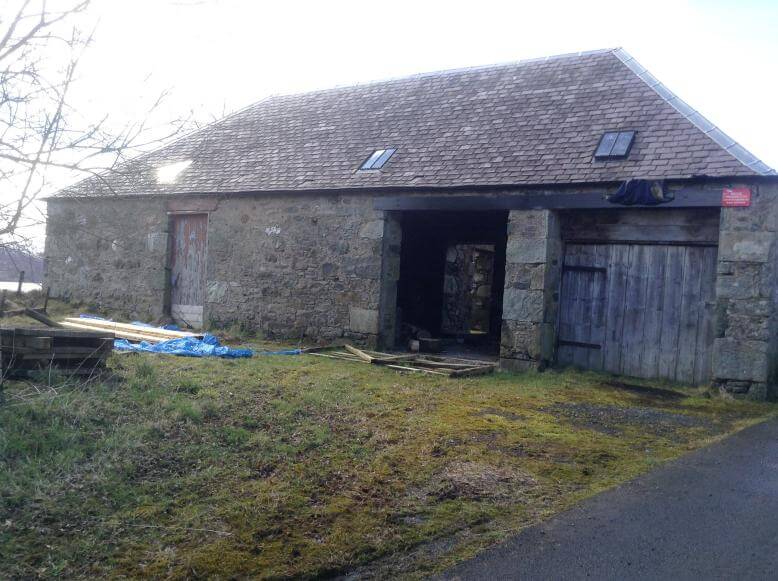 18th century stone farm shed with a 20th-century roof with small skylight windows. Foreground is muddy grass with a paved road on the right