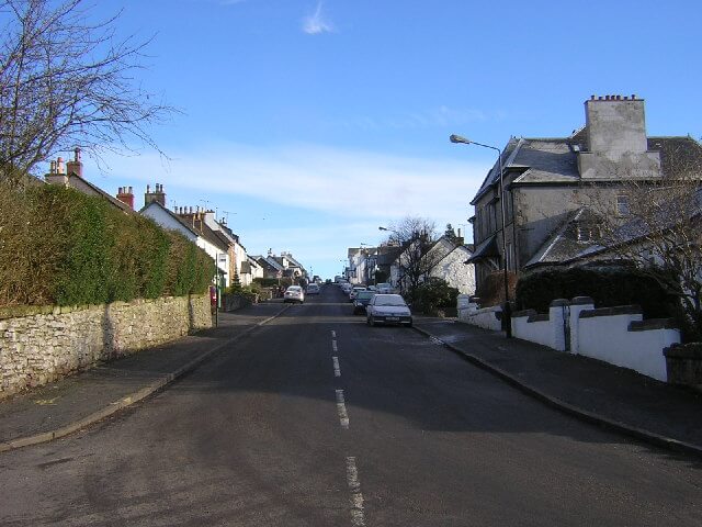 View up a two-lane road of a residential street lined with 19th century houses in a small Scottish town. A few cars are parked along the sides of the street. There is frost visible on the roofs.
