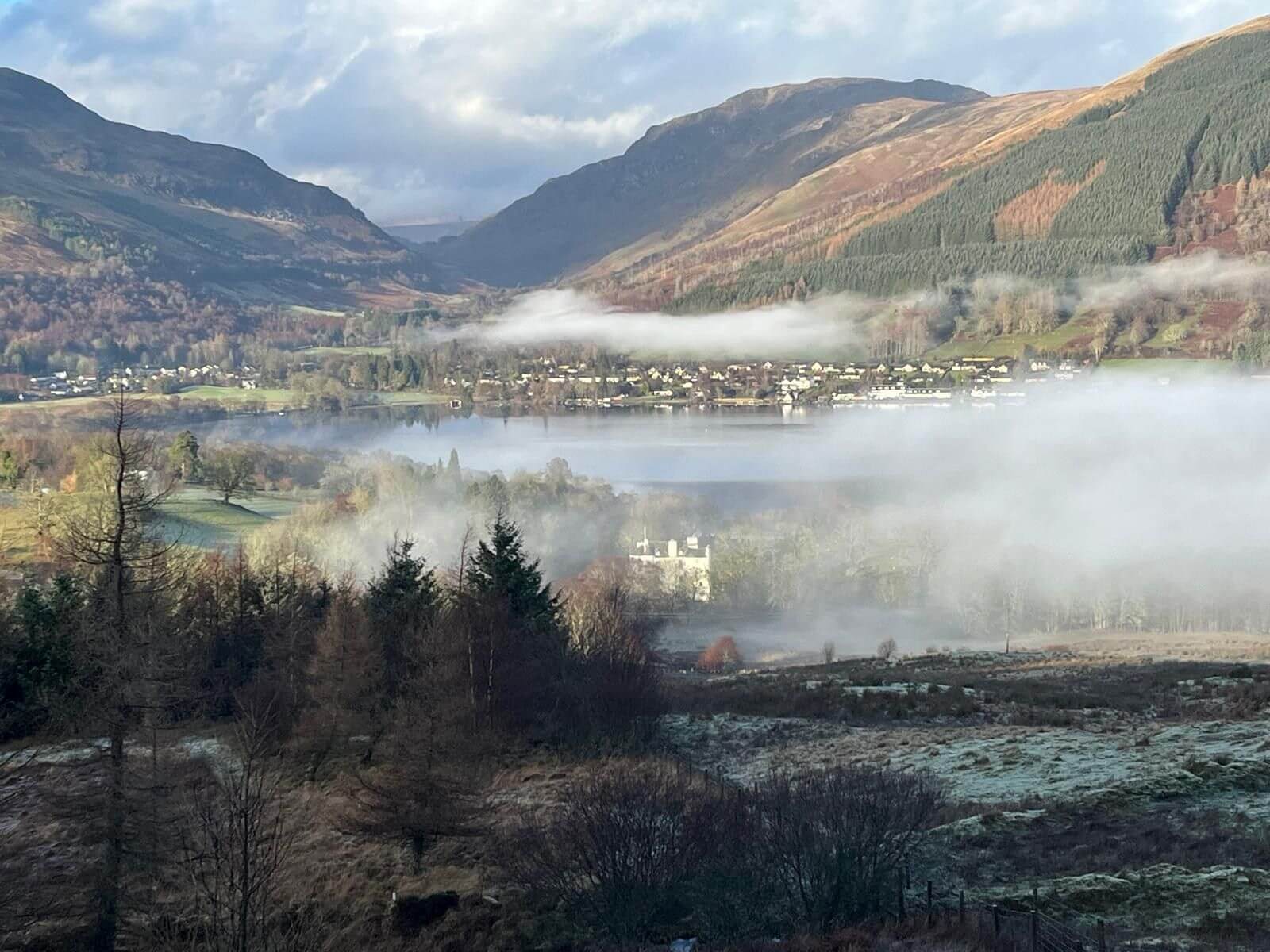 Highland Scottish loch in winter with a mist hanging over the water on an otherwise crisp, sunny day. A mist-shrouded village is visible in the distance. The loch and village are ringed by mountains in the background. A white castle peaks through the mist in the foreground.