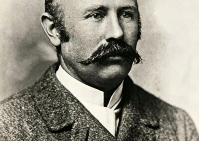 b&w photo, late 1800s, head and torso portrait photo of a slightly balding man who appears to be in his early 50s, with a full and thick curled black moustache. He is wearing a heavy tweed jacket and a white shirt with a stand-up collar.