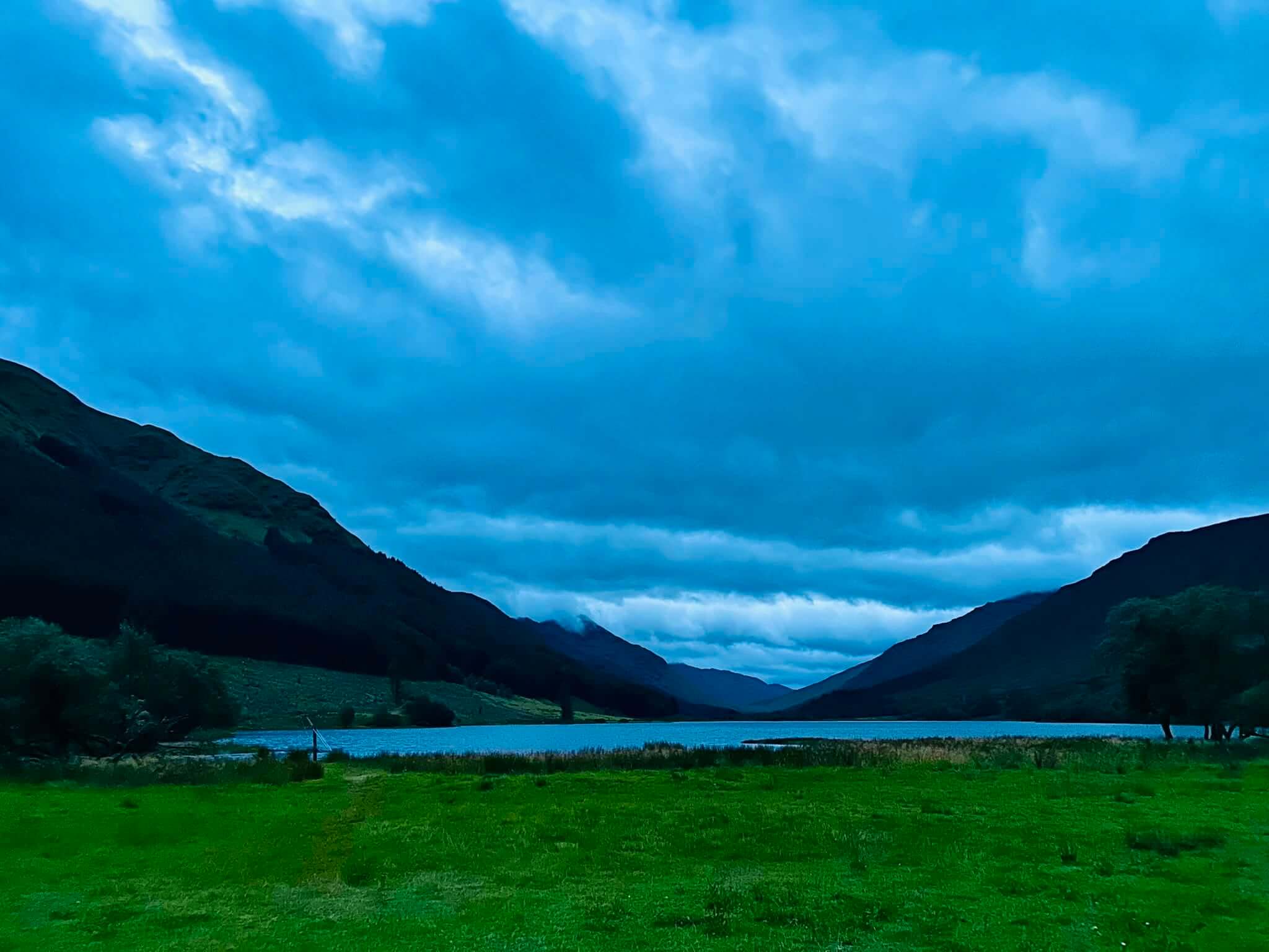 A flat-floored valley in a Highland Scottish glen with mountains rising on either side. Foreground is a field of green grass leading to the shore of a Highland loch that disappears into the distance. The mountains form a 'V' with a very overcast sky. The early morning timing of the photo has turned the overcast clouds a brilliant blue, almost as if the photo was taken through a blue filter.
